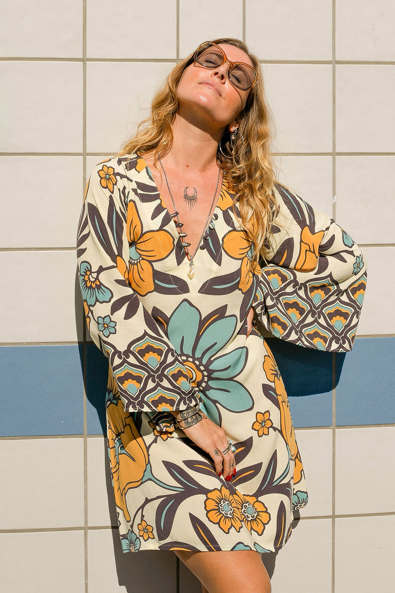 70s vintage inspired boho mini dress with bold retro floral print in blues and mustard on a cream background, bell sleeves and side splits. Made in rayon